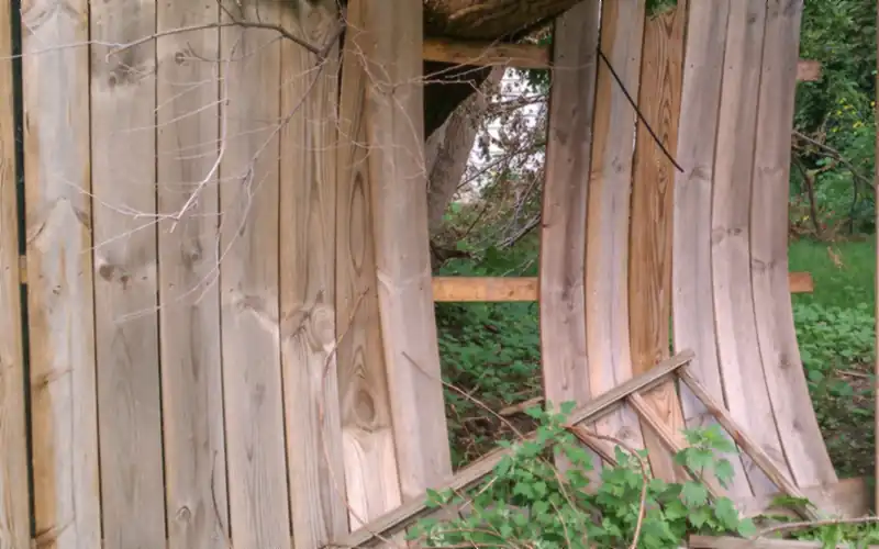 Wood privacy fence broken by a fallen tree, with boards bowed outward and a rail knocked loose